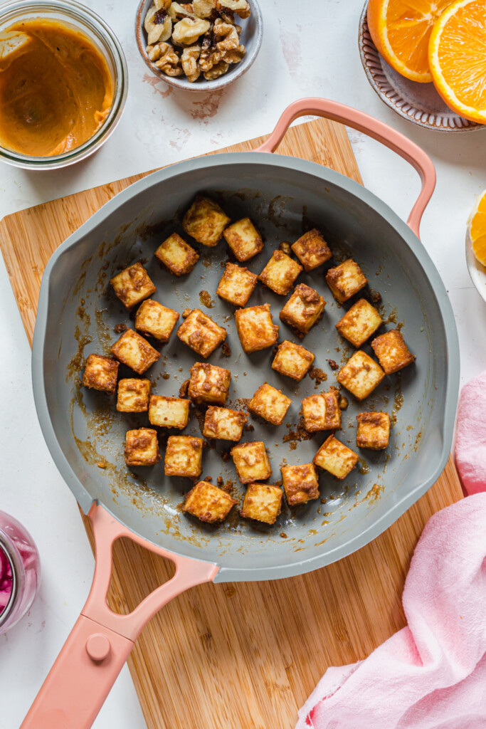 Golden peanut satay fried tofu cubes in a pink and grey pan
