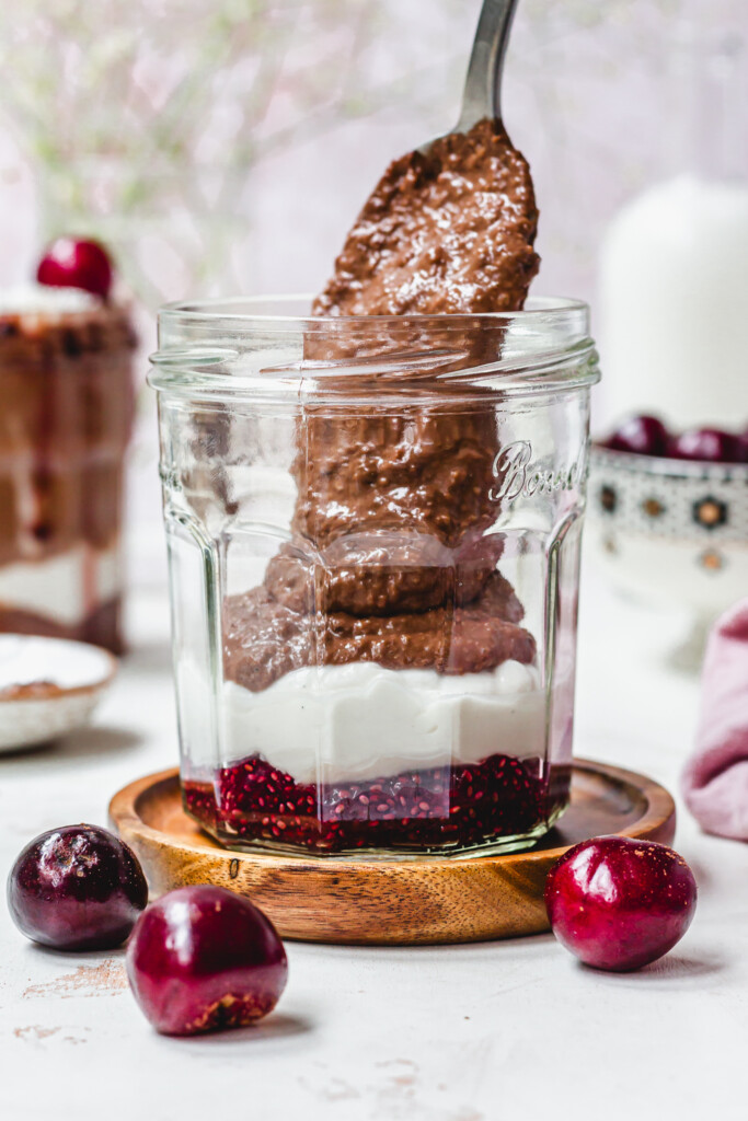 Spooning chocolate oats into a jar with cherry jam and yoghurt