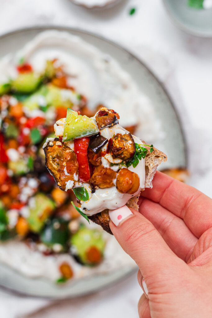 A hand holding a piece of bread topped with Roasted Aubergine and Chickpea Smashed Cucumbers with Whipped Tahini