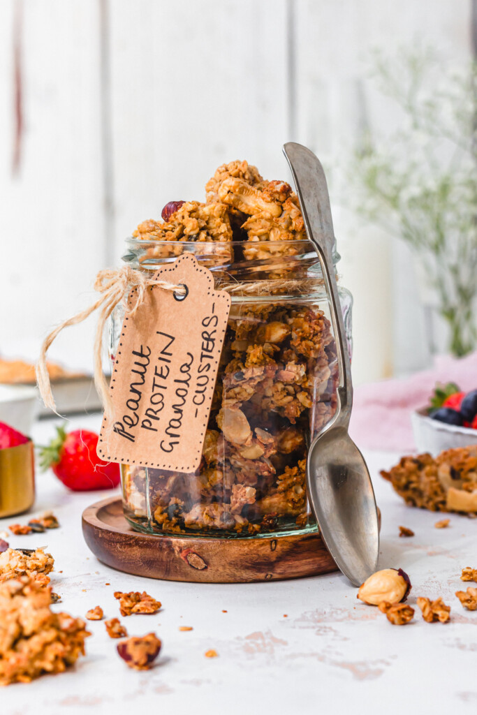 A jar of Peanut Butter Protein Granola Clusters with a spoon and label
